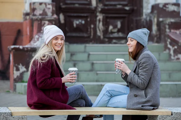 Two sisters relaxing and drinking coffee. Coffee break. Coffee to go.Outdoors fashion portrait of two young beautiful girls drinking coffee. Coffee to go.Two friends relaxing and drinking coffee .Coffee break.Coffee to go