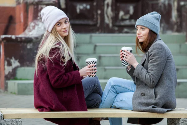 Stylish hipster girl drinking coffee in street.Young girls drinking coffee in city, laughing. Winter weather outdoor. Coffee break. Friends and coffee to go concept.two beautiful long hair girls drinking coffee in the city.