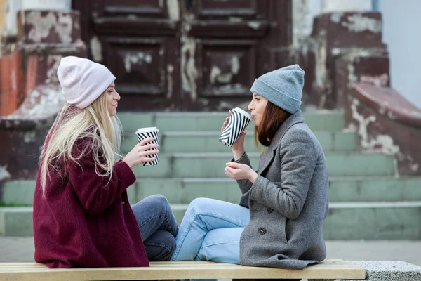 Two sisters relaxing and drinking coffee. Coffee break. Coffee to go.Outdoors fashion portrait of two young beautiful girls drinking coffee. Coffee to go.Two friends relaxing and drinking coffee .Coffee break.Coffee to go