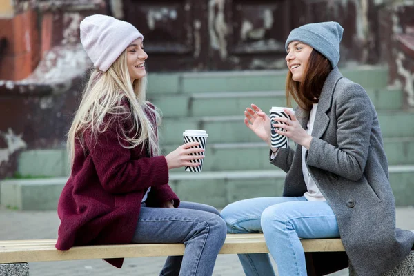 Two sisters relaxing and drinking coffee. Coffee break. Coffee to go.Outdoors fashion portrait of two young beautiful girls drinking coffee. Coffee to go.Two friends relaxing and drinking coffee .Coffee break.Coffee to go