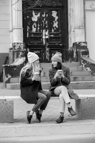 Stylish hipster girl drinking coffee in street.Young girls drinking coffee in city, laughing. Winter weather outdoor. Coffee break. Friends and coffee to go concept.two beautiful long hair girls drinking coffee in the city.