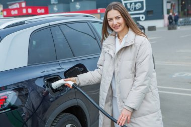 Şehirdeki bir şarj istasyonunda genç bir kadın elektrikli arabasını şarj ediyor. Eko yakıt konsepti. Çevre dostu ulaşım kavramı. Şarj istasyonundan şarj ediliyor..