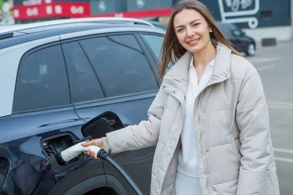 Şehirdeki bir şarj istasyonunda genç bir kadın elektrikli arabasını şarj ediyor. Eko yakıt konsepti. Çevre dostu ulaşım kavramı. Şarj istasyonundan şarj ediliyor..