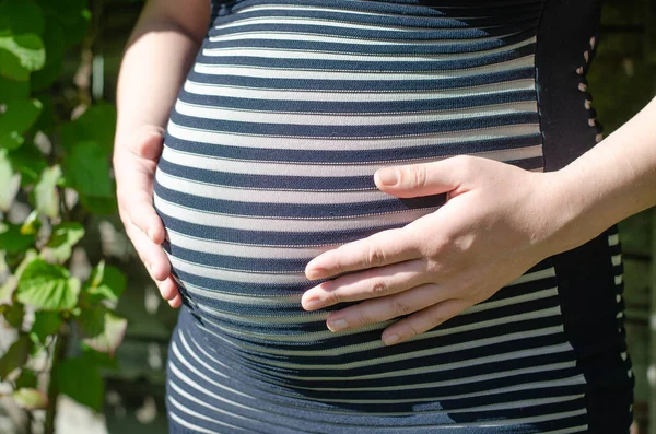 一个穿着条纹衣服的孕妇的照片，腹部特写。手放在肚子上 — 图库照片