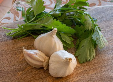 Tahta kesme tahtasının üzerinde üç baş sarımsak, dereotu ve maydanoz resmi.