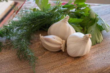 Tahta kesme tahtasının üzerinde üç baş sarımsak, dereotu ve maydanoz resmi.
