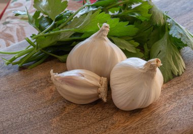 Tahta kesme tahtasının üzerinde üç baş sarımsak, dereotu ve maydanoz resmi.