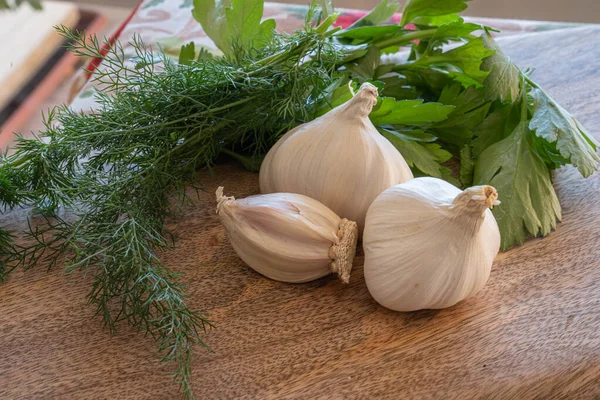 Tahta kesme tahtasının üzerinde üç baş sarımsak, dereotu ve maydanoz resmi.