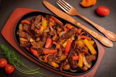 Koyu bir arka planda dekorasyon olarak eski çatal bıçak ve sebzelerle ahşap bir tezgahta, dökme demir bir tavada fajitos resmi