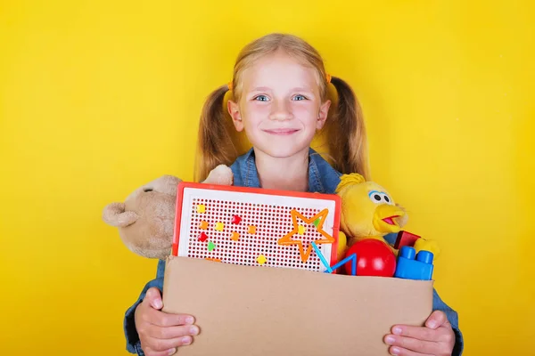 Blond little girl holding a box with toys on yellow background. Donation concept.. — Stock Photo, Image