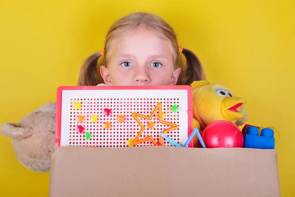 Sarışın küçük kız sarı arka planda oyuncaklar ile bir kutu tutan. Bağış kavramı.. — Stok fotoğraf