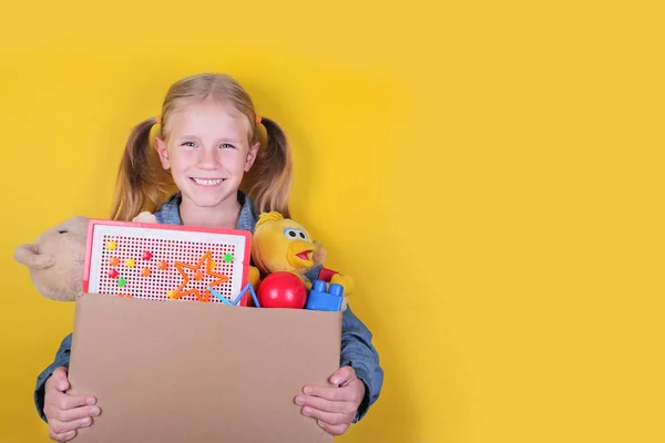 Blond little girl holding a box with toys on yellow background. Donation concept.. — Stock Photo, Image