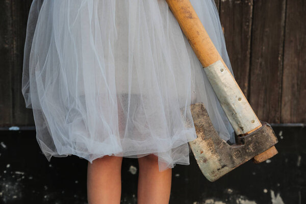 Smiling blond girl holding an axe in hands on wooden background. Halloween celebration,  horror concept.
