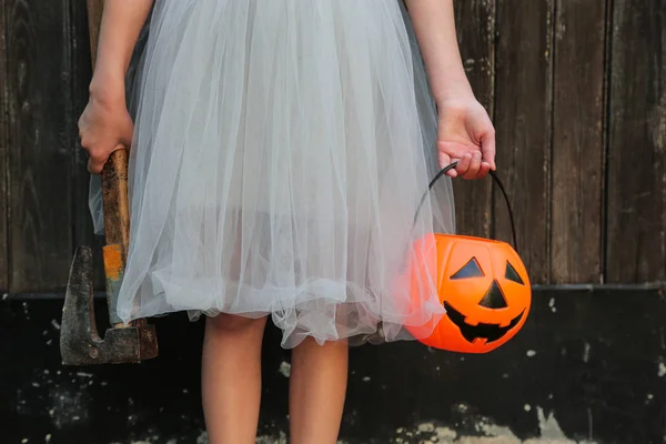 Chica rubia sonriente sosteniendo calabaza de Halloween jack o 'lantern y un hacha en las manos sobre fondo de madera. Halloween celebración, concepto de horror . —  Fotos de Stock