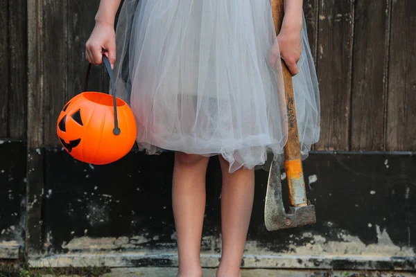 Glimlachend blond meisje houdt Halloween pompoen Jack O'Lantern en een bijl in handen op houten achtergrond. Halloween viering, horror concept. — Stockfoto