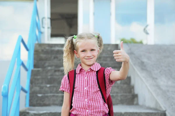 Back to school. Beautiful blond schoolgirl is showing thumb up gesture with red backpack and books outside the primary school , education concept. — Stock Photo, Image