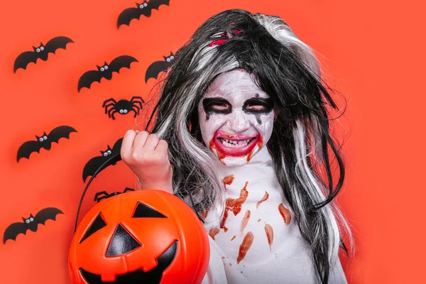 Concepto de horror de Halloween. Linda niña asustadiza en traje de zombi monstruo con calabaza sobre fondo naranja . —  Fotos de Stock