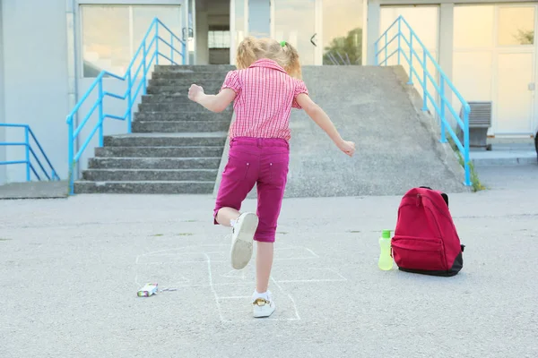 Back to school. Beautiful blond schoolgirl playing hopscotch drawn with colorful chalk on asphalt outside the primary school , education concept. — Stock Photo, Image