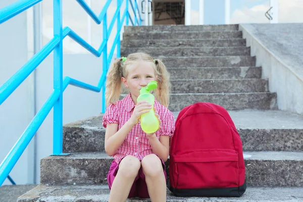 Back to school. Beautiful blond schoolgirl drinking water from a bottle with red backpack and books outside the primary school , education concept. — Stock Photo, Image