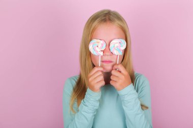 Komik, gülümseyen sarışın kız gözlerini pembe arka planda lolipoplarla kaplıyor..