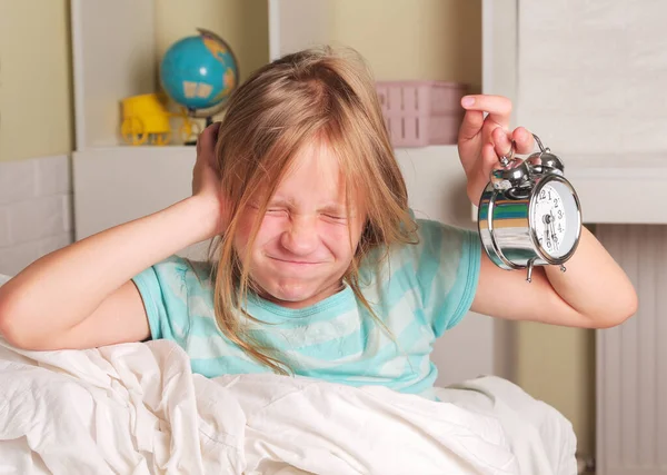 School girl hates waking up early in the morning. Blond girl holding alarm clock , yawning in bed covers ear with hand