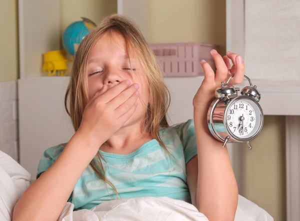 School girl hates waking up early in the morning. Blond girl holding alarm clock , yawning in bed covers ear with hand