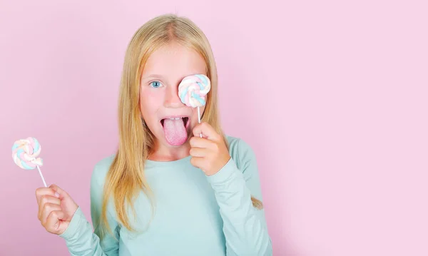 Grappig kind meisje bedek ogen met lolly 's marshmallow en tonen de tong op roze achtergrond. — Stockfoto