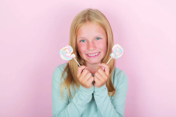 Engraçado sorrindo menina loira criança com pirulitos marshmallow no fundo rosa. — Fotografia de Stock