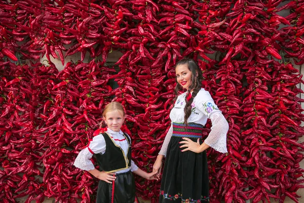 Hagyományos szerb balkáni ruhába öltözött lányok, nemzeti népviselet. Pózol közelében tétel piros paprika paprika paprika lóg a falon a ház. . — Stock Fotó