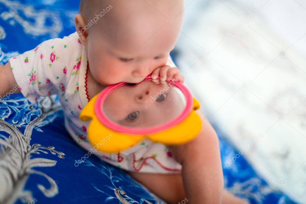 Adorable and cute baby girl sits on the bed and plays with toys that is children mirror. Small child is holding flower toy in the mouth and looking through mirror.