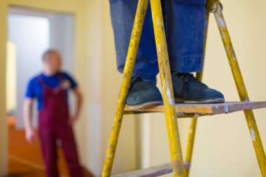 Mavi iş giysisi ve ahşap merdiven üzerinde siyah ayakkabı ile alt bacaklar. İşçi Alçıpan yapar veya Alçıpan alçı duvarlar daire için yenileme, yenileme, revizyon, uzantısı, restorasyon ve rekonstrüksiyon, henüz tamamlanmamıştır