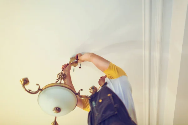 Worker in workwear is installing a chandelier and connects it  to electricity on the ceiling from ladder in apartment is under construction, remodeling, renovation, overhaul, extension, restoration an — Stock Photo, Image