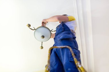 Worker in workwear is installing a chandelier and connects it  to electricity on the ceiling from ladder in apartment is under construction, remodeling, renovation, overhaul, extension, restoration an clipart