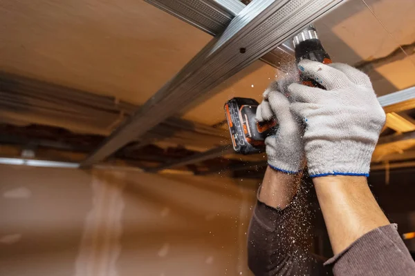 Работник в защитных перчатках сверлит перфоратором потолок для установки металлических рам в строящейся квартире, которая находится в стадии строительства, реконструкции, реконструкции, капитального ремонта, расширения, реставрации и — стоковое фото