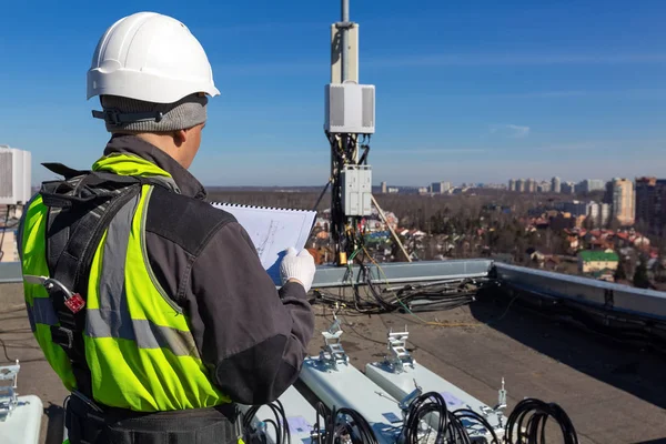 El escalador industrial profesional en casco y uniforme lee documentación técnica y antenas de bandas GSM DCS UMTS LTE, unidades de radio al aire libre en el techo. Proceso de mejora —  Fotos de Stock