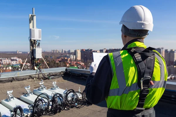 El escalador industrial profesional en casco y uniforme lee documentación técnica y antenas de bandas GSM DCS UMTS LTE, unidades de radio al aire libre en el techo. Proceso de mejora — Foto de Stock
