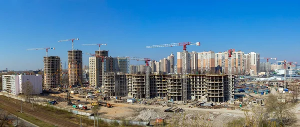 Panorama of large construction site including several cranes working on a building complex, workers, construction gear, tools and equipment, trucks and clear blue sky as background.