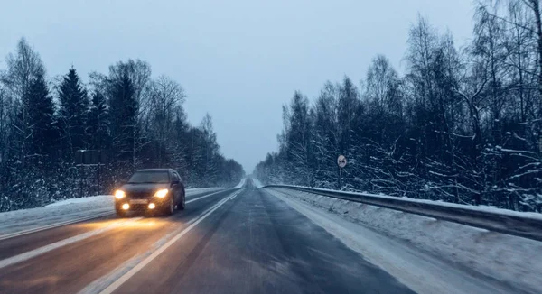 雪が飛んでいるとき、夕暮れの雪の嵐の中で冬の道路にヘッドライトを付けて車を運転します。冬に悪い視界と危険な条件で運転の概念。動きを持つ画像 — ストック写真