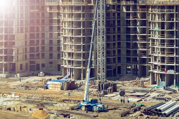 Large construction site including several cranes working on a building complex, workers, construction gear, tools and equipment, tractor and apartments without walls and windows are as background.