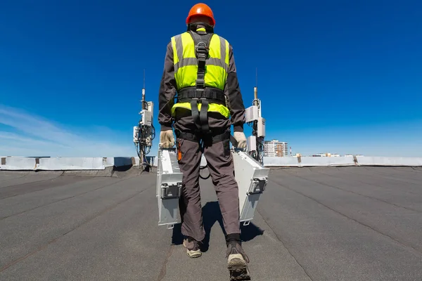 Alpinista industrial profissional em capacete e uniforme detém equipamentos de telecomunicações em sua mão e antenas de bandas GSM DCS UMTS LTE, unidades de rádio ao ar livre, fibras ópticas, cabos de alimentação são instalados — Fotografia de Stock