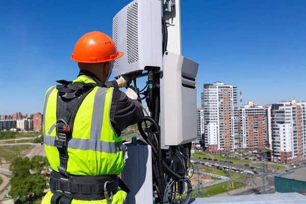 Ingegnere delle telecomunicazioni in casco e uniforme installa apparecchiature di telecomunicazione in mano e antenne di bande GSM DCS UMTS LTE, unità radio all'aperto, fibre ottiche, cavi di alimentazione sono installati — Foto Stock