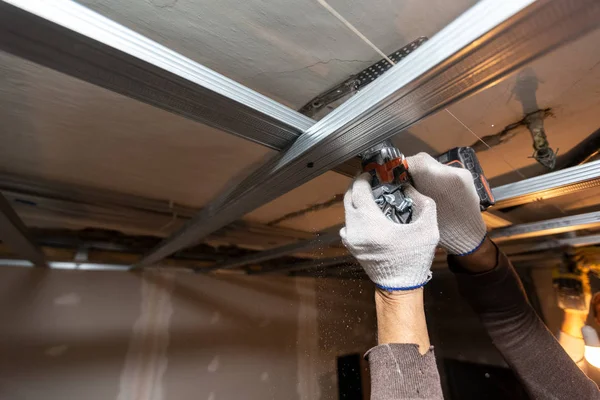 Работник в защитных перчатках сверлит перфоратором потолок для установки металлических рам в строящейся квартире, которая находится в стадии строительства, реконструкции, реконструкции, капитального ремонта, расширения, реставрации и — стоковое фото