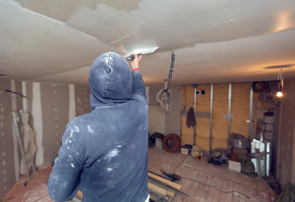 Worker is working with palette-knife for flattening the ceiling from wooden platform in room of apartment is inder construction, remodeling, renovation, overhaul, extension, restoration and — Stock Photo, Image