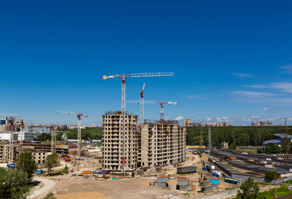 Large construction site including several cranes working on a building complex, workers, construction gear, tools and equipment, trucks and  clear blue sky as background.