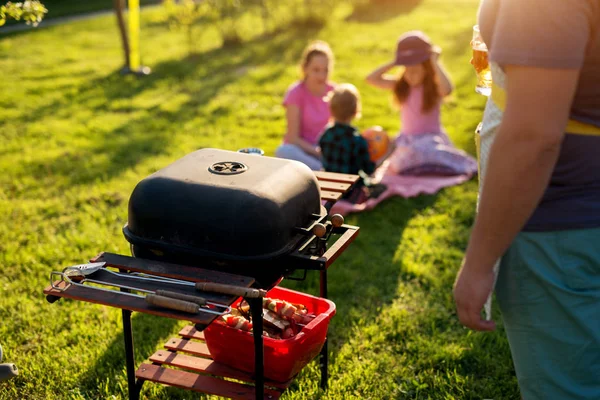 stock image Man is about to use a vintage old clean grill with meat and vegetables already ready beneath it while kids are chatting and sitting on the blanket that has been put on grass.