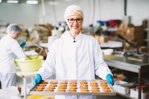 たてを保持している若い幸せな女性労働者は食品工場でブリキのクッキーを焼きました — ストック写真