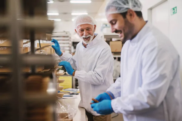 Picture of two male food factory employees in sterile clothes packing fresh made cookies and having fun.