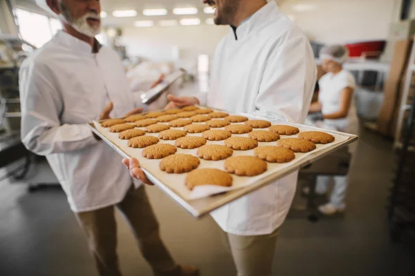 食品工場の大きな皿の新鮮なクッキーをクローズ アップ 無菌服バック グラウンドで話の つの男性従業員の画像をぼやけ 一人の男がトレイを持っています — ストック写真
