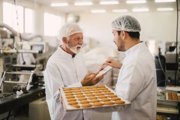 笑顔でお返事の食品工場で無菌服で 人の従業員の写真 以前はタブレットを保持 生産工程をチェック 若い男がトレイがいっぱいの新鮮なクッキーを保持しています — ストック写真