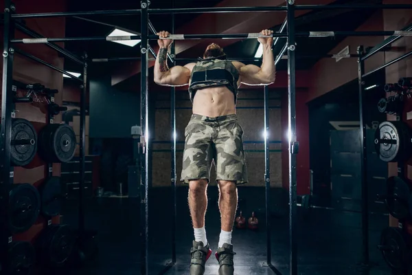 Muscular caucasian bearded man doing pull-ups in military style weighted vest in crossfit gym. Weight plates, kettlebells, barbell and crossfit tires in background.